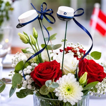 Blomsterpind med blå studenterhue placeret i blomsterdekoration med røde roser og hvide blomster som bordpynt til studenterfest.