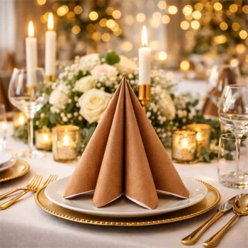 Elegant festbord med kobberfarvede airlaid servietter, gyldent bestik, stearinlys og blomster i varm stemningsbelysning