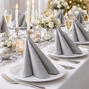 Elegant festbord med sølvfarvede Airlaid servietter, hvide tallerkener, lys og blomster i stilfuld borddækning