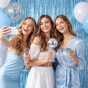 Tre smilende kvinder tager selfie foran en lyseblå glimmerforhæng fotovæg med balloner og festpynt.
