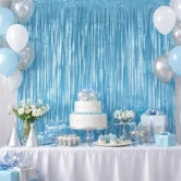 Festlig baggrund med lyseblåt glimmerforhæng bag et dækket kagebord med hvid lagkage, blå balloner og festpynt.